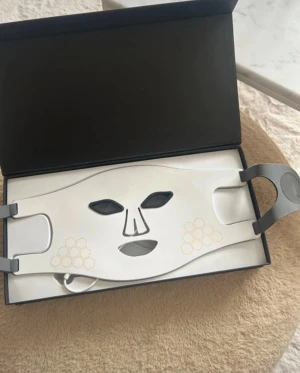 LED ansiktsmask med fjärrkontroll - Säljer en vit LED ansiktsmask. Masken har röda, blå och lila LED-lampor och kommer med en vit fjärrkontroll för att justera inställningar.