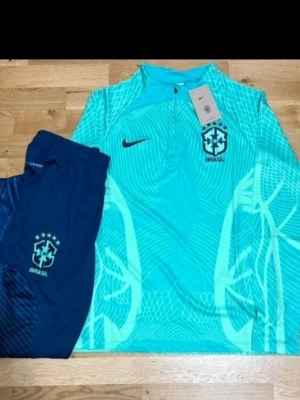 Nike Brazil football kit - Brasilien träningsset från Nike med långärmad tröja och shorts, men inga skor finns med. Tröjan är turkos med grafiskt mönster och shorts är marinblå med Brasilien-logga.