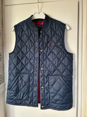 Mörkblå quiltad dunväst från Polo Ralph Lauren - Snygg mörkblå quiltad dunväst från Polo Ralph Lauren med klassisk röd logga på bröstet. Västen har tryckknappar i guld, två stora fickor framtill och en hög krage. Perfekt att slänga över en hoodie eller tröja för extra värme.
