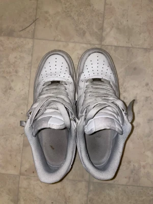 Nike Air Force 1 vita sneakers - Fräscha vita Nike Air Force 1 sneakers med klassisk design och perforerad tå för extra ventilation. Skorna har snörning, platt sula och rund tå. Tillverkade i skinn med mjukt foder och tydlig Nike-logga på sidan. Perfekta för dig som gillar streetstyle och vill ha en ikonisk sneaker.