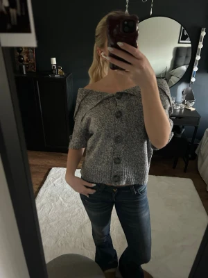 Grå offshoulder stickad kofta - Säljer en gråmelerad stickad kofta med offshoulder-design och stora knappar framtill. Koftan har korta ärmar och ribbad nederkant, perfekt för dig som gillar en chill och trendig look. Passar snyggt till jeans eller kjol.