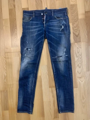 Dsquared2 jeans - Säljer ett par blå jeans från Dsquared2 med coola slitningar och färgstänk på benen. Jeansen har klassisk femficksdesign, knappgylf och brunt läderpatch bak. Passformen är smal och materialet är robust denim. Perfekt för dig som gillar en edgy streetstil. Själv fallet autentiska. 