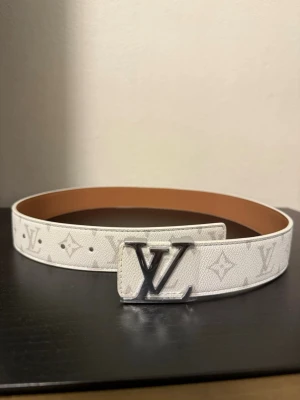 Bälte - Snyggt vitt bälte från klassiskt monogrammönster och stor spänne i silver. Bältet är gjort i präglat skinn med ljusbrun insida och har en bred, stilren form som verkligen sticker ut. Perfekt för dig som gillar exklusiva accessoarer.