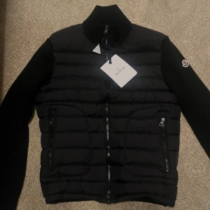 Moncler Cardigan - Svart Cardigan från Moncler med stickade ärmar och quiltad kropp. Jackan har två dragkedjeförsedda fickor, hög krage och Moncler-logga på ärmen. Materialmix av polyester och stickad textil ger en snygg kontrast. Perfekt för dig som gillar stilren design. Pris går att diskutera. Jackan är hel ny endast provad.