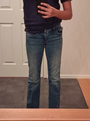 Blå bootcut jeans med kors på bakfickorna - Säljer ett par blå bootcut jeans med snygga kors och paljetter på bakfickorna. Jeansen har låg midja och klassisk femficksdesign. Materialet är jeans med lätt tvättad look och passformen är normal. Perfekt för dig som gillar lite extra detaljer på dina jeans. Byxorna är uppsydda av sömmerska