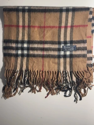 Rutig beige Burberrys kashmir halsduk - Säljer en klassisk halsduk  från Burberrys i 100% kashmir med det ikoniska rutiga mönstret i beige, svart, vitt och röda linjer. Västen har fransar nedtill och en mjuk, lyxig känsla. Perfekt för dig som gillar tidlös design och vill sticka ut. (10C)