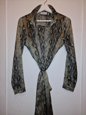 Sally Snake klänning  från Neo Noir - Snygg klänning från Neo Noir med coolt ormmönster i grönt och svart. Klänningen har knytband i midjan för en smickrande siluett. Använd 1 gång. En utav knapparna är borta och är lätt att fixa till, vilket inte syns då det är dubbla tyg över knapparna. Se sista bilden. 