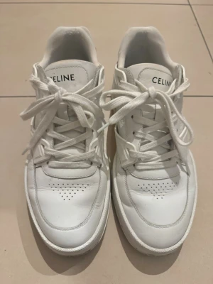 Vita sneakers från Celine strl 42 - Säljer ett par vita Celine sneakers i storlek 42. Skorna har klassisk rund tå, perforerade detaljer på tån och tydlig Celine-logga både på plösen och hälen. Tillverkade i skinn med snörning och platt sula för en clean och stilren look. Dom är äkta men har inget kvitto eller kartong kvar