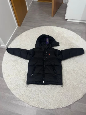 Svart pufferjacka från Polo Ralph Lauren - Svart polo Ralph jacka. Man kan se stygnen längst upp vid luvan ungefär därför kan vi ta de dm och diskutera om pris. Den är helt ny