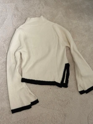 Vit polotröja med svarta detaljer - Säljer en ribbad polotröja i vit färg med svarta kanter på ärmslut och nederkant. Tröjan har lång ärm och en liten slits på sidan, vilket ger en cool touch. Perfekt för dig som gillar stilrena och trendiga plagg.