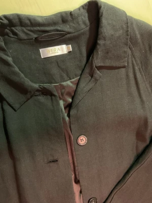 Svart lång kappa från Wera - Svart kappa från Wera med klassisk krage och knäppning framtill. Lång modell med diskreta fickor i sidorna. Yttertyg i viskos, tencel och lin, fodrad med polyester. Perfekt för dig som gillar stilrena och tidlösa plagg.