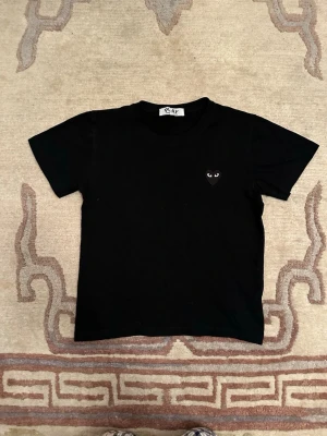  Comme des Garçons Play - Svart t-shirt från Comme des Garçons Play med klassisk rund hals och korta ärmar. På bröstet sitter det ikoniska hjärta med ögon, broderat i svart. T-shirten är gjord i mjuk bomull och har en clean, stilren vibe.