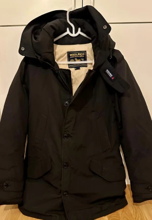 Woolrich jacka - storlek/M i jättebra skick!  - Woolrich jackor är det som främst kännetecknar varumärket, med fokus på vinterjackor skapade för att klara arktisk kyla. Ord pris över 9000 kr mitt pris är endast 2499 kr.  Svart parkajacka från Woolrich med huva, stora fickor och knappar framtill. Jackan är tillverkad i Ramar Cloth, en mix - beige insida. Perfekt för kalla dagar och har en clean, klassisk look./ pris kan diskuteras.
