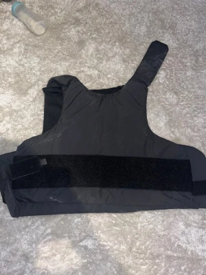 Svart skyddsväst XL med kardborre - Säljer en svart skyddsväst i storlek XL med breda axelband och kraftig kardborre framtill. Västen har en stilren design och är tillverkad i slitstarkt syntetmaterial. Perfekt för dig som vill ha en funktionell och enkel väst med justerbar passform.