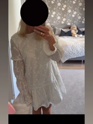 Studentklänning från NA-KD - Superfin vit studentklänning från NA-KD med blommigt broderat mönster och volangdetaljer. Klänningen har långa, transparenta ärmar och en rund halsringning. Endast använd 1 gång så mkt fint skick. Den är öppen i ryggen