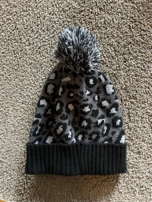 Grå leopardmönstrad stickad mössa - Säljer en stickad mössa i grå och svart med leopardmönster och en stor fluffig tofs på toppen. Mössan har bred ribbad kant nedtill och är perfekt för kalla dagar. Den har en cool och trendig vibe som passar till streetwear.