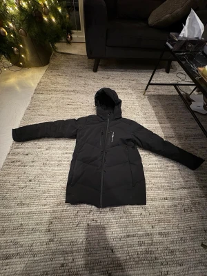 Svart dunjacka från sail Racing - Svart dunjacka från sail Racing med huva och flera praktiska fickor med dragkedja. Jackan är quiltad och har en clean look med logga på bröstet. Fodrad med Allied Feather + Down, 500 FP. Perfekt för kalla dagar och har lång ärm.