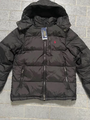 Svart pufferjacka från Polo Ralph Lauren - Svart pufferjacka från Polo Ralph Lauren med huva och dragkedja framtill. Jackan har det klassiska röda Polo-logot broderat på bröstet och är tillverkad i polyester. Perfekt för kalla dagar med sin quiltade design och långa ärmar.