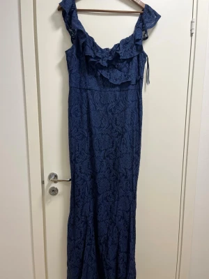 Marinblå långklänning med volang - Säljer en marinblå långklänning med vackert blommönster i spets och volangdetalj vid axlarna. Klänningen har en elegant, figurnära passform och är perfekt för dig som vill ha något extra till speciella tillfällen.