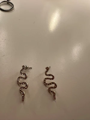 Glittriga örhängen i ormform - Säljer ett par snygga örhängen formade som ormar, täckta med glittriga stenar. De är silvriga och har en cool, slingrande design som verkligen sticker ut. Perfekta för dig som vill ha något unikt och trendigt i smyckeslådan.
