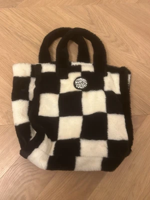 Rutig fluffig handväska Cosmic Youth - Säljer en supermjuk och fluffig handväska från Cosmic Youth med svartvita rutor. Väskan har två rejäla handtag och ett coolt broderat märke framtill. Perfekt för dig som gillar statement-accessoarer och vill sticka ut med din look.