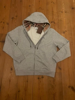 Grå Burberry zip-hoodie - Ljusgrå zip-hoodie från Burberry i mjukt och bekvämt material. Tröjan har hel dragkedja, huva med Burberrys ikoniska rutiga foder samt ribbade muddar vid ärmslut och nederkant. Praktiska fickor framtill. Helt ny med originalprislapp kvar, aldrig använd. 
