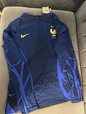 Ingen skobild – ingen skobeskrivning - Bilderna visar en blå Nike-tröja med franska landslagets emblem, inte skor. Ladda upp bilder på skor för att få en detaljerad skobeskrivning och taxonomier.