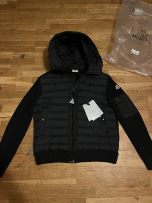 Svart Moncler Cardigan med huva - Säljer den för att den är liten på mig och har inte använt denna så den är sprillans ny! Passar dig som har storlek S och M. Svart Cardigan jacka från Moncler med stickade ärmar, huva och dragkedja framtill. Jackan har Moncler-logga på ärmen, praktisk ficka med dragkedja på ena ärmen och två sidofickor. Materialmix av dun och stickad polyester ger en schysst look.