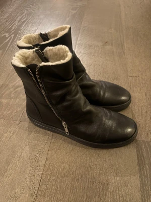 Svarta fodrade boots med dragkedja - Säljer ett par svarta boots i skinn med rund tå och platt sula. Skorna har vit, fluffig foder på insidan och dragkedja på sidan för enkel på- och avtagning. Perfekta för kalla dagar och har en stilren look som passar till många outfits.