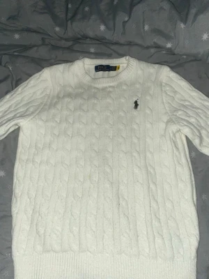 Vit stickad tröja från Polo Ralph Lauren - Säljer en vit stickad tröja från Polo Ralph Lauren med klassiskt kabelstickat mönster och rund hals. Tröjan har långärmade och ribbade muddar vid ärmslut och nederkant. Den ikoniska Polo-loggan är broderad på bröstet.