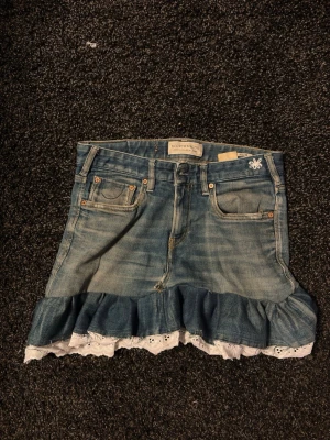 Jeanskjol med volang från Scotch & Soda - Säljer en själv sydd ljusblå jeanskjol från Scotch & Soda med volangkant och vit spetsdetalj längst ner. Kjolen har klassiska jeansfickor fram och bak, samt knapp och dragkedja i midjan. Perfekt för dig som gillar en söt och avslappnad stil.