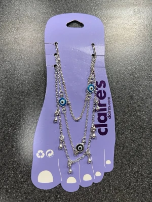 Silverigt fot armband onda ögat från Claire's - Säljer en snygg fo tarmband i silverfärg från Claire's med flera kedjor och hängande droppar och det onda ögat. Armbandet har blå och svart evil eye-detaljer som ger en cool vibe. Perfekt accessoar för att lyfta din stil och ge lite edge till dina sneakers eller sandaler.