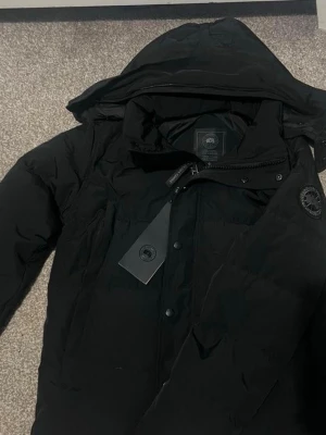 Svart dunjacka från Canada Goose - Säljer en svart dunjacka från Canada Goose med huva och snygga detaljer som dragkedja och knappar framtill. Jackan har en klassisk patch på ärmen och är perfekt för kalla dagar. Stilsäker och varm, passar dig som vill ha både funktion och stil.