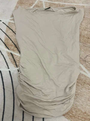 Topp Gina tricot  - Säljer en beige tubtopp med snygg rynkdetalj på sidan. Toppen är kortärmad och har en enkel, clean design som är lätt att matcha med olika outfits. Materialet känns mjukt och stretchigt, perfekt för varma dagar eller när du vill ha en chill look.