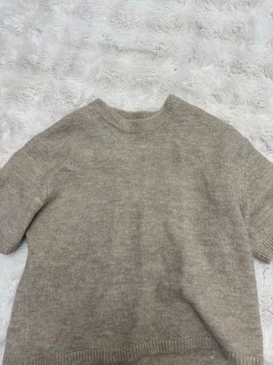 Beige stickad tröja från Gina Tricot - Mysig brun/beige stickad tröja från Gina Tricot i ullblandning. Tröjan har korta ärmar, rund hals och ribbade kanter vid hals, ärmslut och nederkant. Perfekt för lager-på-lager under kyliga dagar.