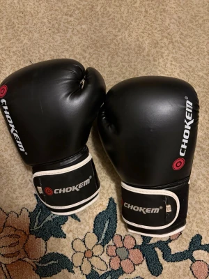 Svarta boxningshandskar Chokem 12oz - Säljer ett par svarta boxningshandskar från Chokem i storlek 12oz. Handskarna har vit kant och röd logga, med kardborreband för enkel justering. Tillverkade i syntetmaterial med mjuk stoppning för bra komfort under träning.