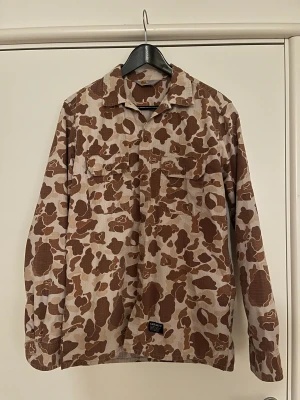 Camouflage overshirt från Carhartt - Säljer en snygg overshirt från Carhartt med orange, brunt och beige camouflage-mönster. Jackan har två bröstfickor med knappar och klassisk krage. Jag har använt jackan ett fåtal gånger. Hör av dig vid frågor eller funderingar!
