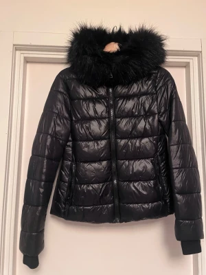 Zara Svart pufferjacka med fin fuskpäls  - Svart pufferjacka från Zara med glansig yta och fin, fluffig fuskpäls runt huvan. Jackan har dragkedja framtill, två sidofickor med dragkedja och ribbade muddar vid ärmsluten. Mycket bra skick! Storlek S. Upplagd på flera säljsidor, först till kvarn. 