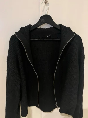 Svart stickad hoodie med dragkedja - Svart stickad hoodie från 157 med grov ribbad struktur och hel dragkedja framtill. Tröjan har huva och långa ärmar, perfekt för chill dagar. Materialet är mjukt och ger en avslappnad loose passform. Snygg att matcha med jeans eller joggers.