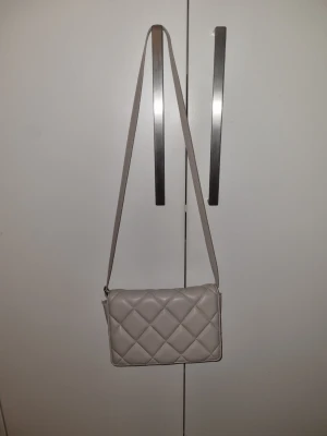 Beige quiltad axelväska från H&M - Säljer en beige axelväska från H&M med quiltad design och lång axelrem. Väskan har ett magnetlås och flera fack inuti för enkel organisering. Perfekt storlek för mobil, plånbok och andra småsaker. Stilren och enkel att matcha med olika outfits.