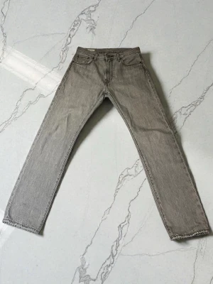 Levi's 551 grå jeans W29 L32 - Grå jeans från Levi's modell 551 med klassisk femficksdesign och raka ben. Jeansen har dragkedja och knappstängning, samt detaljerad läderpatch bak i midjan. Materialet är slitstark bomullsdenim med Water<Less-teknologi. Jeansen är lite slitna på höger framficka (se bild 5) annars är de i mycket bra skick.