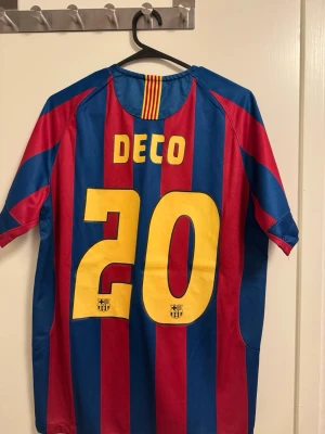 Barcelona Deco 20 matchtröja - Säljer en klassisk FC Barcelona matchtröja med Deco och nummer 20 på ryggen. Tröjan har breda blå och röda ränder, gult Nike-märke och klubbemblem på bröstet. Tillverkad i polyester, perfekt för fotbollsfans som vill sticka ut med en ikonisk retrostil.