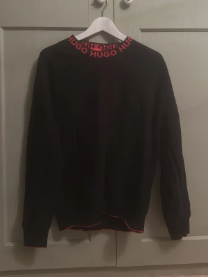Svart tröja från Hugo med röd logga - Original pris 1600kr. Svart långärmad tröja från Hugo med röd logga runt halsen och röda detaljer vid muddar och nederkant. Tröjan har en klassisk passform och är perfekt för dig som gillar stilrena plagg med en twist. Snygg att matcha med jeans eller chinos.