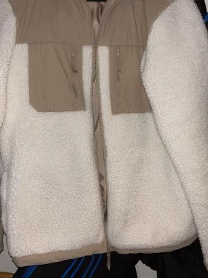 Beige och vit teddyjacka med dragkedja - Säljer en mysig teddyjacka i beige och vitt med två bröstfickor med dragkedja. Jackan har en loose passform och är tillverkad i fleece och syntetmaterial. Perfekt för dig som gillar chill och comfy stil.