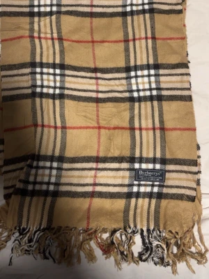 Rutig beige scarf från Burberry - Säljer en klassisk rutig scarf från Burberry  i beige, svart, vit och röd. Scarfen är tillverkad i 100% lammull och har fransar längst ner. Den har det ikoniska Burberry-mönstret och är perfekt för dig som gillar tidlös stil.