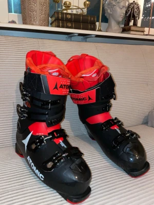 Atomic Hawx 130s Magna pjäxor - Säljer ett par Atomic Hawx 130s Magna pjäxor i svart och rött med robusta spännen och bred kardborre upptill. Insidan är mjukt fodrad i röd-orange ton för extra komfort. Perfekt för dig som vill ha stabilitet och kontroll i backen. Grym design och coola detaljer.