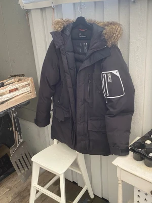 Svart Peak Performance parkas med päls - Peak Performance parkas i svart med huva och snygg pälsdetalj. Jackan har flera fickor, dragkedjor och en vit Gore-Tex patch på ärmen. Perfekt för kalla dagar med sin långa och quiltade design. R&D-logga på huvan och reflekterande detalj nedtill.