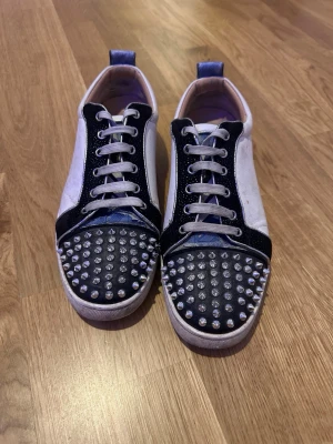 Loubs  - Unika sneakers från Christian Louboutin med svart och vit ovandel, blå detaljer på hälen och röd sula. Tåpartiet är täckt av silvriga nitar för en edgy look. Skorna har snörning och platt sula, tillverkade i skinn och textil. Perfekt för dig som vill sticka ut.