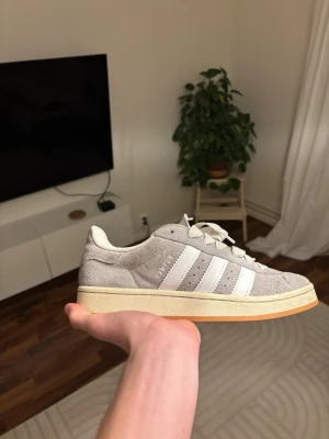 Adidas Campus grå sneakers - Säljer ett par Adidas Campus sneakers i ljusgrå mocka med vita klassiska stripes och snörning. Skorna har en platt sula i beige och gummi, samt rund tå. Perfekta för dig som gillar retrostil och vill ha något som funkar till jeans eller shorts.
