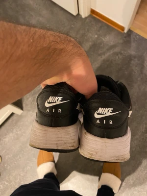 Svarta Nike Air sneakers med vit sula - Säljer ett par svarta Nike Air sneakers med vit sula och vit swoosh på sidan. Skorna har snörning och är tillverkade i en kombination av mesh och skinn för extra komfort och stil. Klassisk Nike Air-dämpning i sulan och rund tå. Perfekt för dig som gillar sportig streetstil.